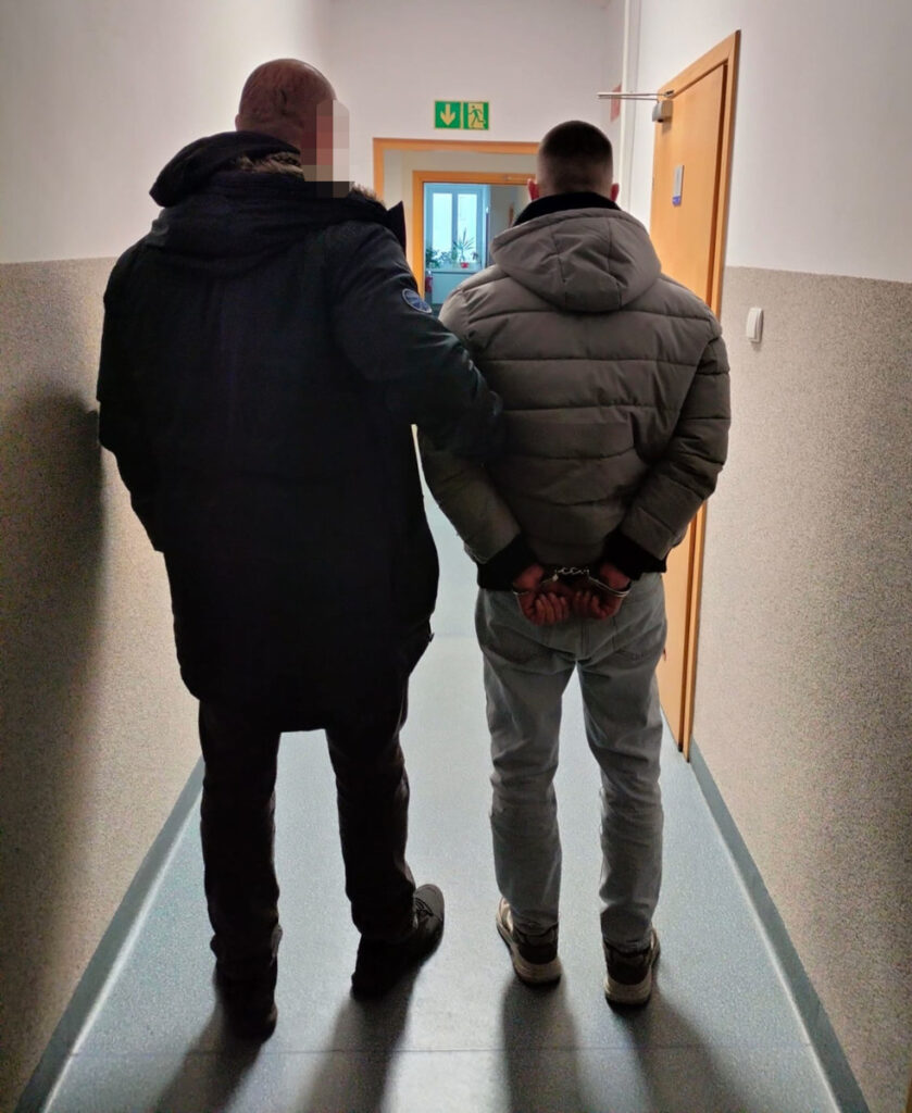 Mężczyzna w ciemnej kurtce prowadzi osobę w szarej kurtce z rękami związanymi z tyłu, idących korytarzem.