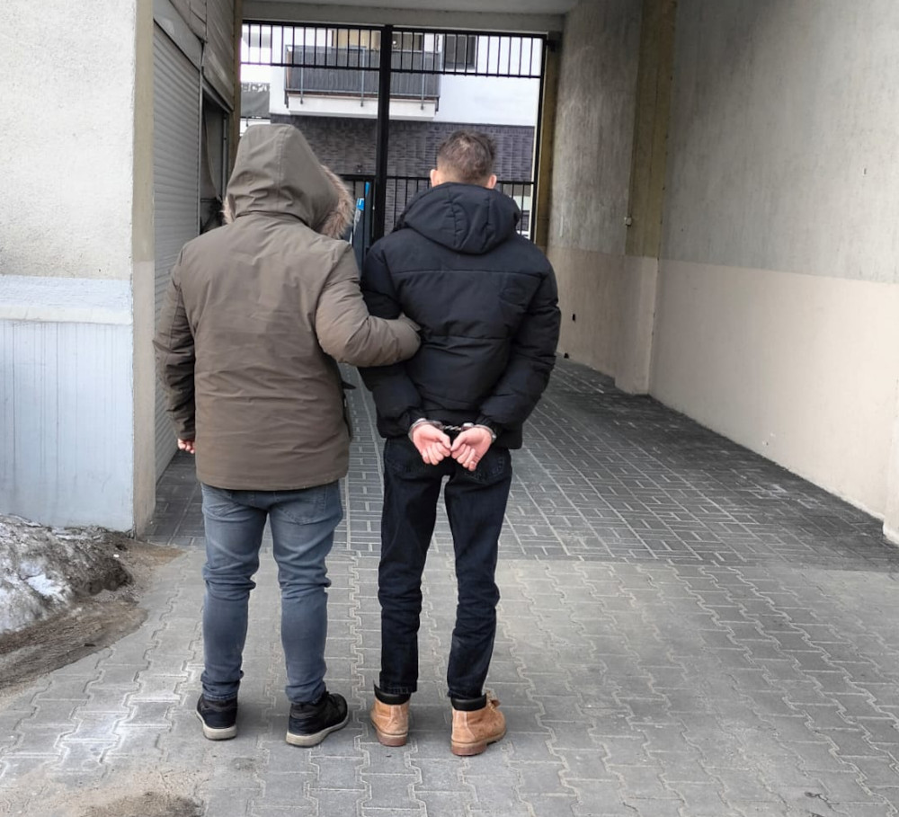 Mężczyzna w kurtce prowadzi drugiego mężczyznę z rękami związanymi z tyłu w wąskim przejściu.