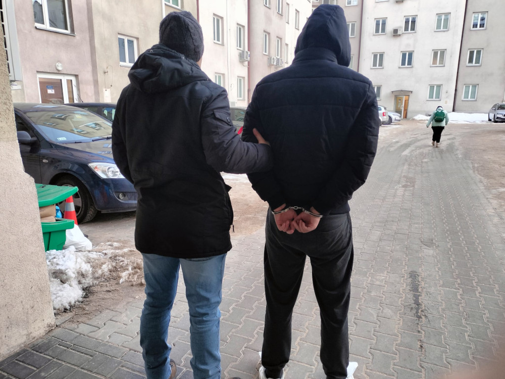 Mężczyzna w kajdankach prowadzony przez funkcjonariusza w podwórku z blokami mieszkalnymi.