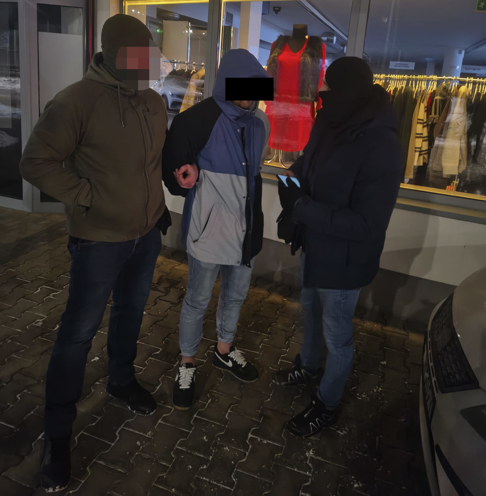 Dwaj mężczyźni w ciemnych ubraniach rozmawiają z osobą w jasnej kurtce na tle wystawy sklepowej.