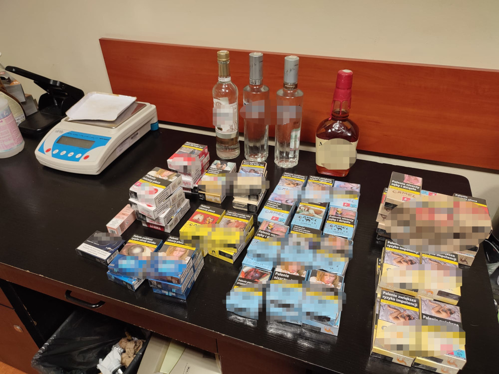 Na czarnym blacie leżą opakowania papierosów różnych marek, butelki alkoholu oraz waga elektroniczna.