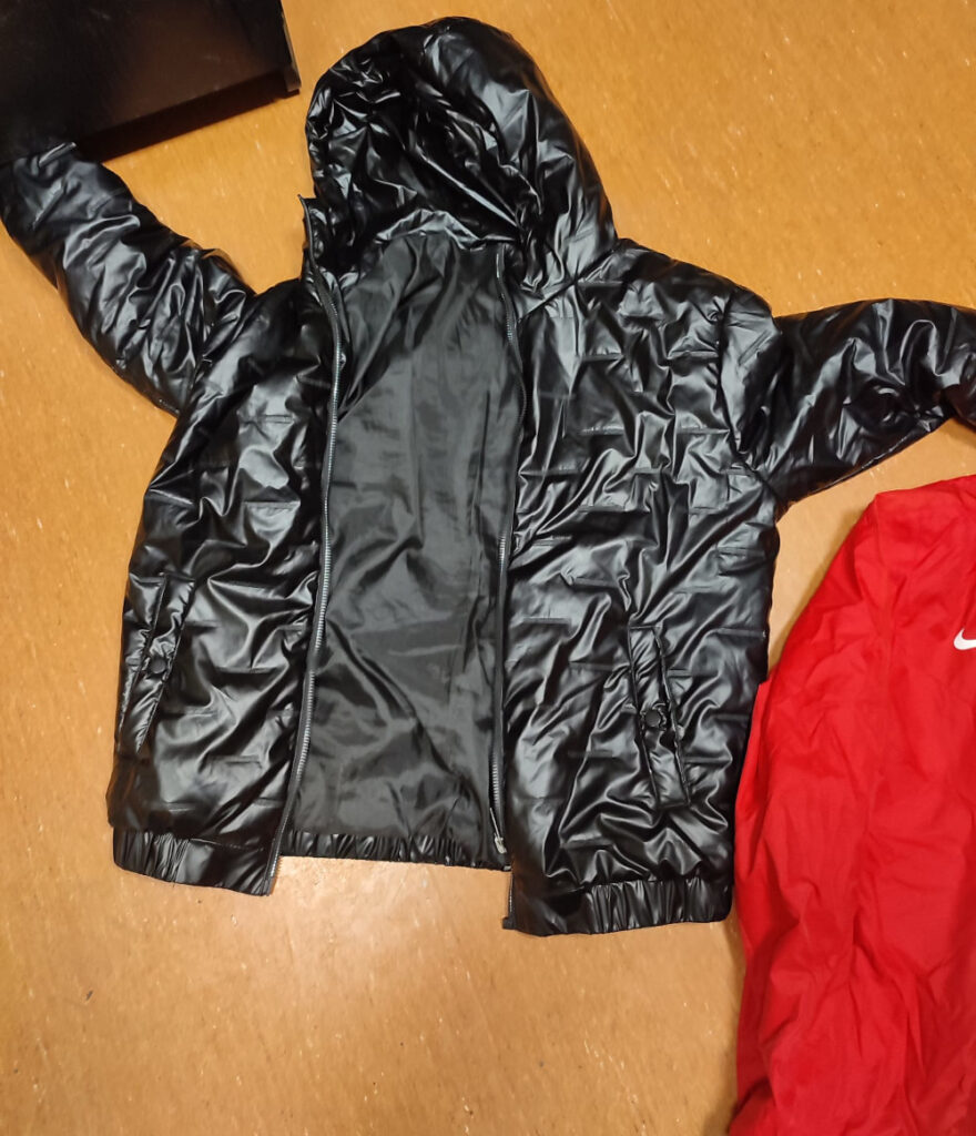 Czarna kurtka puchowa z kapturem leży na drewnianej podłodze obok czerwonej kurtki marki Nike.