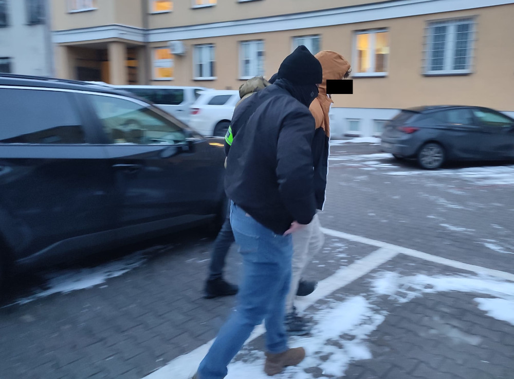 Mężczyzna w ciemnej kurtce prowadzi osobę w pomarańczowej bluzie przez parking w zimowej scenerii.