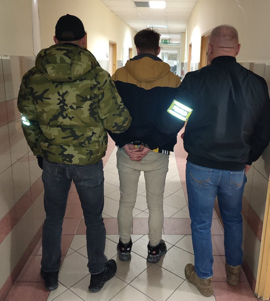 Trzech mężczyzn idących korytarzem, jeden z nich z rękami związanymi z tyłu, pozostali trzymają go za ramiona.