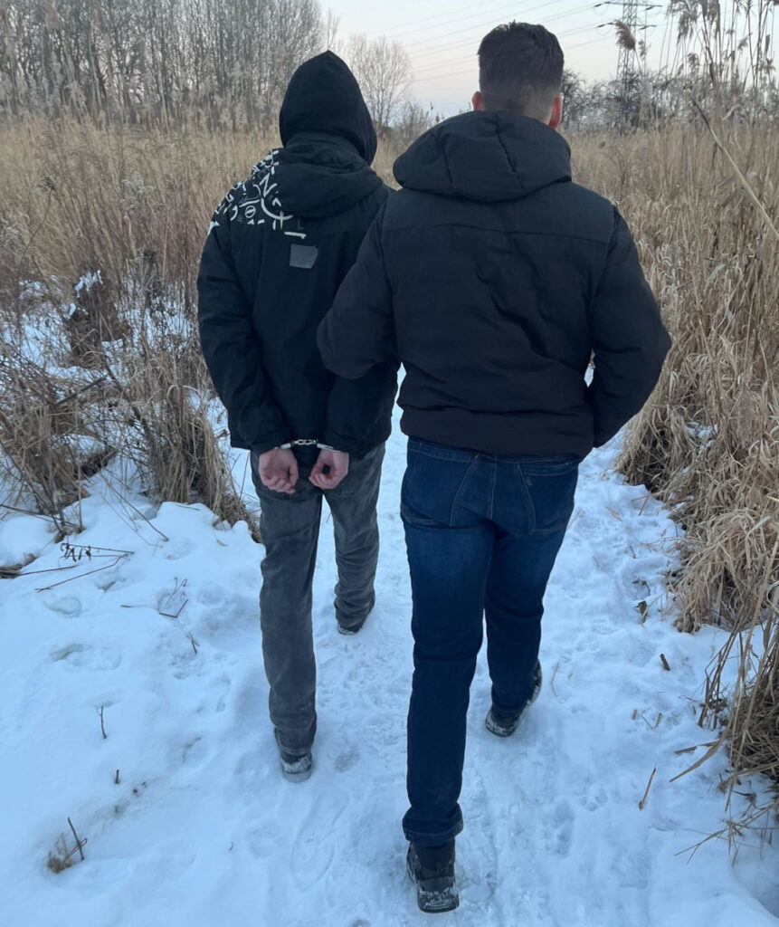 Mężczyzna w kajdankach prowadzony przez drugiego mężczyznę w zimowym otoczeniu, na śnieżnej ścieżce.