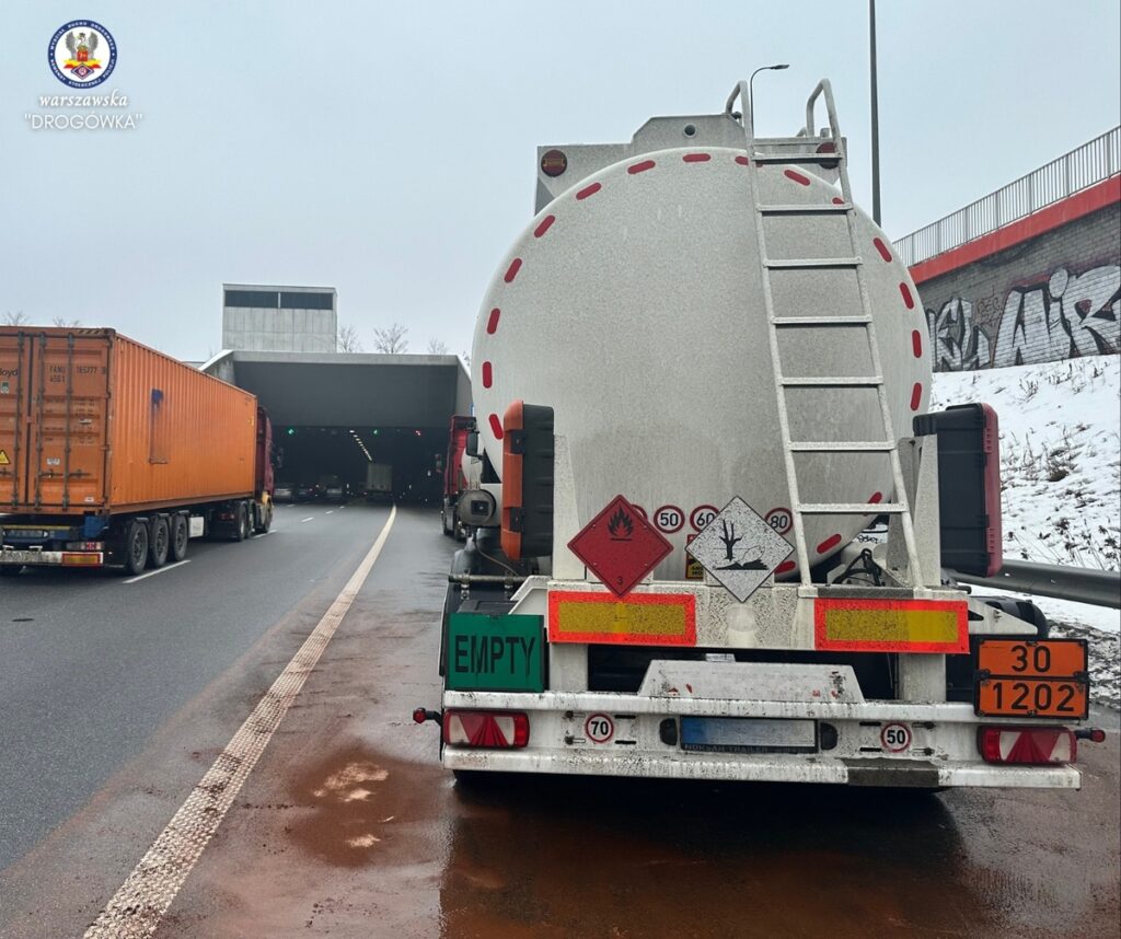 Czarny ciągnik siodłowy z naczepą transportującą kontener oraz cysternę na drodze, w tle wjazd do tunelu.