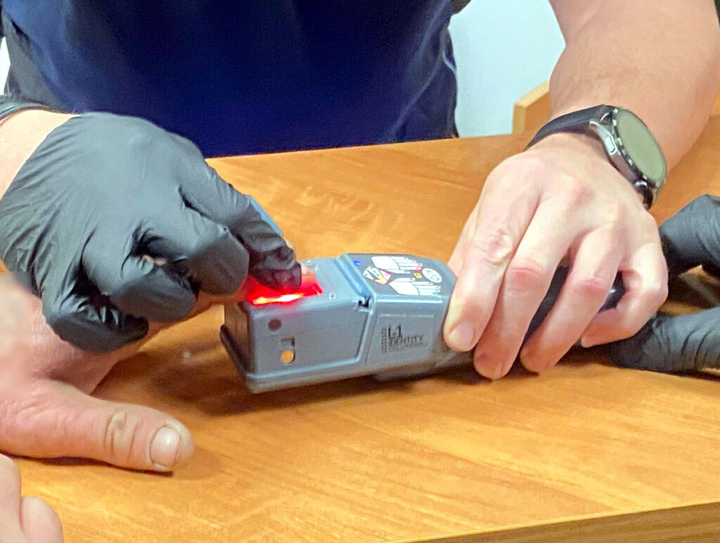 Mężczyzna w rękawiczkach obsługujący urządzenie z czerwoną diodą na drewnianym stole.