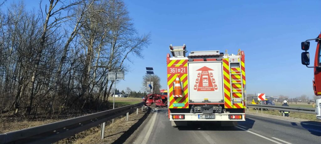 Czerwony wóz strażacki stoi na poboczu drogi obok zniszczonego samochodu, w otoczeniu drzew i znaków drogowych.