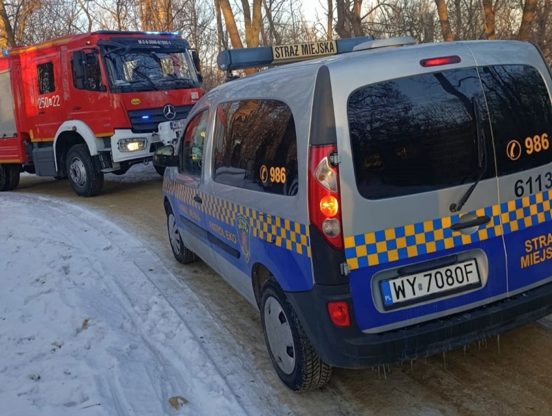 Straż miejska w niebiesko-żółtym samochodzie stoi na zaśnieżonej drodze obok czerwonego wozu strażackiego.
