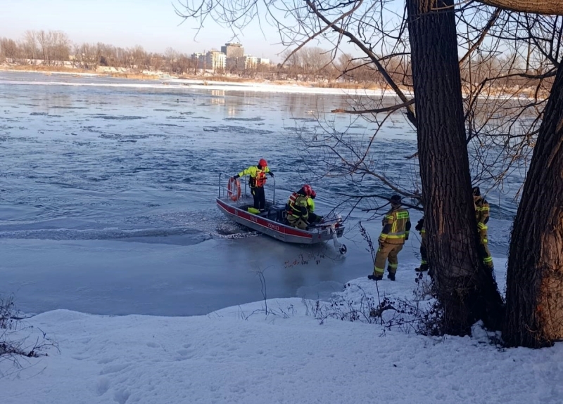 Ratownicy w kamizelkach na łodzi motorowej przeszukują zamarzniętą rzekę w zimowym otoczeniu.
