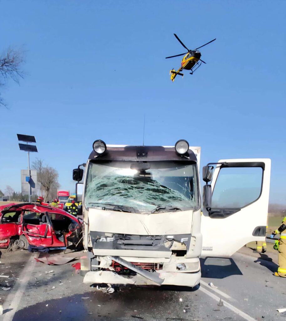 W miejscu wypadku drogowego, rozbity ciężarowy pojazd stoi na jezdni obok uszkodzonego samochodu osobowego, nad nimi krąży śmigłowiec ratunkowy.