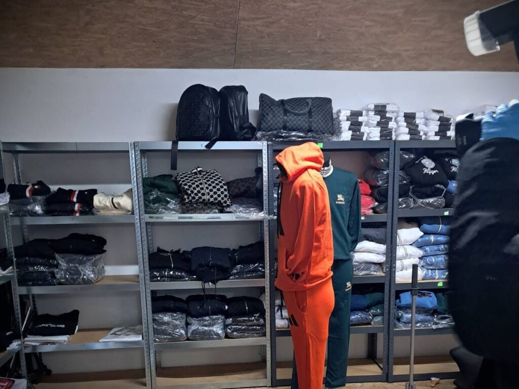 Manekin w pomarańczowym dresie stoi obok regałów z odzieżą w różnych kolorach, w tle torby i zrolowane ubrania.