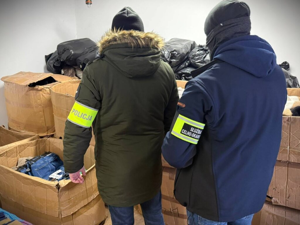 Funkcjonariusze w odzieży służbowej analizują zawartość kartonów z odzieżą w magazynie.