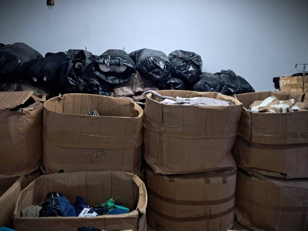 W pomieszczeniu znajdują się duże kartony z ubraniami, obok których leżą czarne worki pełne odzieży.
