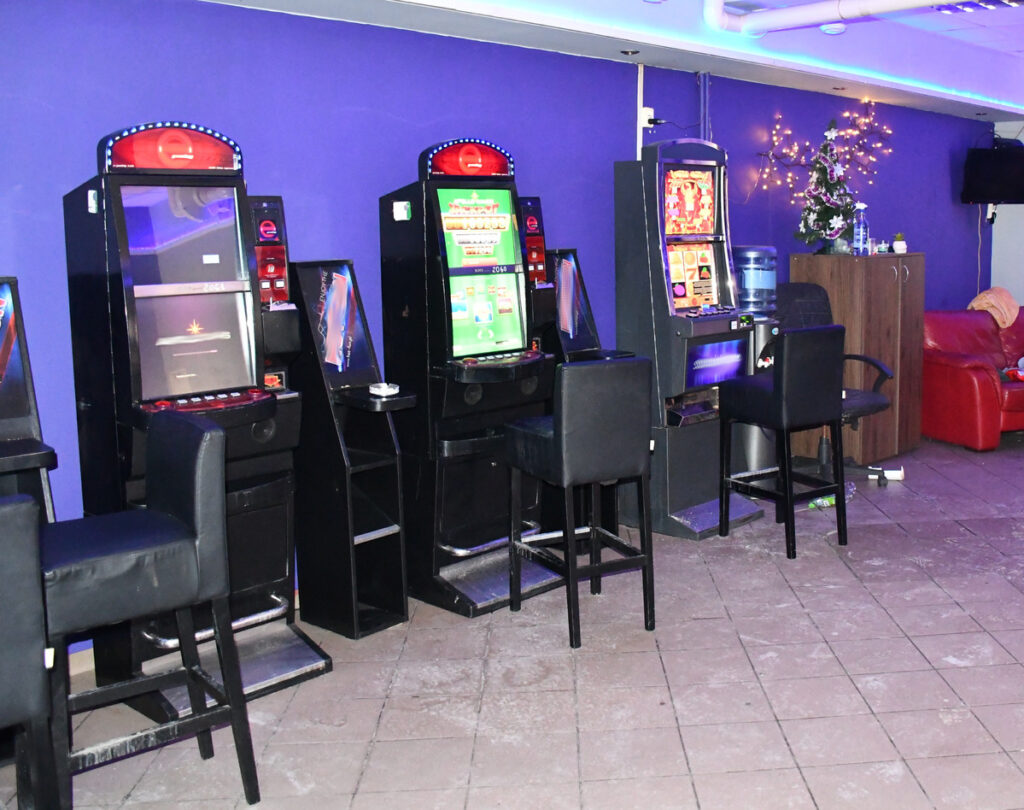 Automaty do gier w ciemnym pomieszczeniu z fioletowymi ścianami, obok czarne krzesła barowe i stół z wodą.