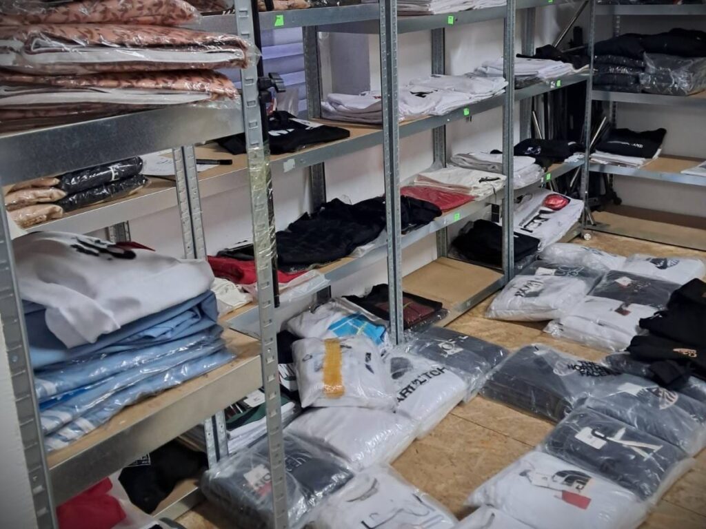 Półki z odzieżą w magazynie, na których znajdują się złożone koszulki i bluzy, obok leżące zapakowane ubrania.