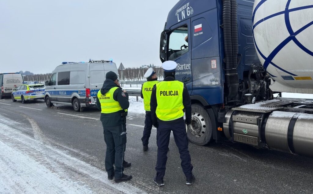 Policjanci w kamizelkach odblaskowych kontrolują kierowcę ciężarówki na zaśnieżonej drodze.