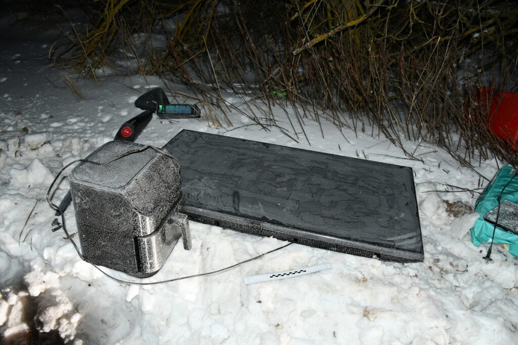 Zamarznięty komputer i ekran leżą na zaśnieżonym terenie, obok widać narzędzia oraz gałęzie krzewów.