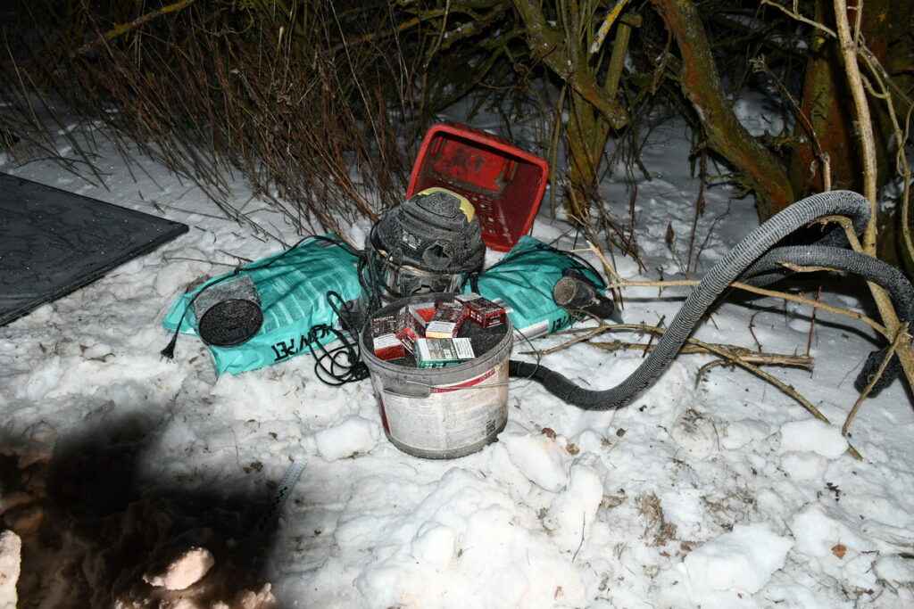 Na śniegu leży odkurzacz obok pojemnika z paczkami papierosów i zielonym workiem, otoczony gałęziami.