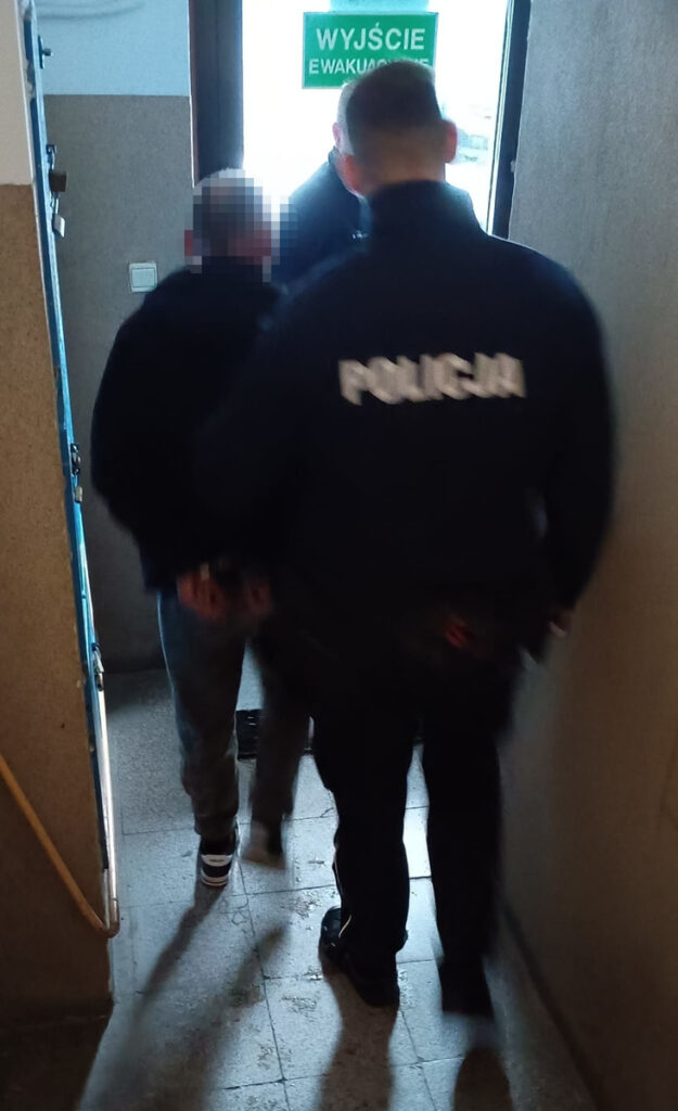 Mężczyzna w kajdankach prowadzony przez policjanta w korytarzu budynku, z wyjściem ewakuacyjnym w tle.
