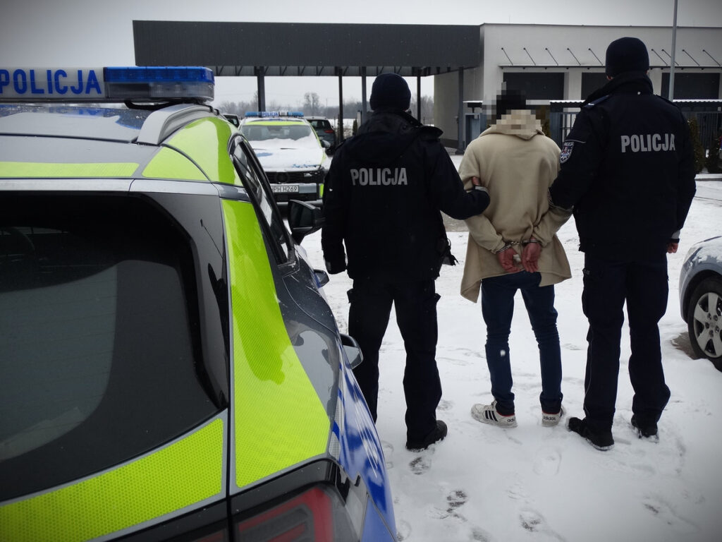 Mężczyzna w kajdankach prowadzony przez dwóch policjantów na śniegu, obok policyjny radiowóz.