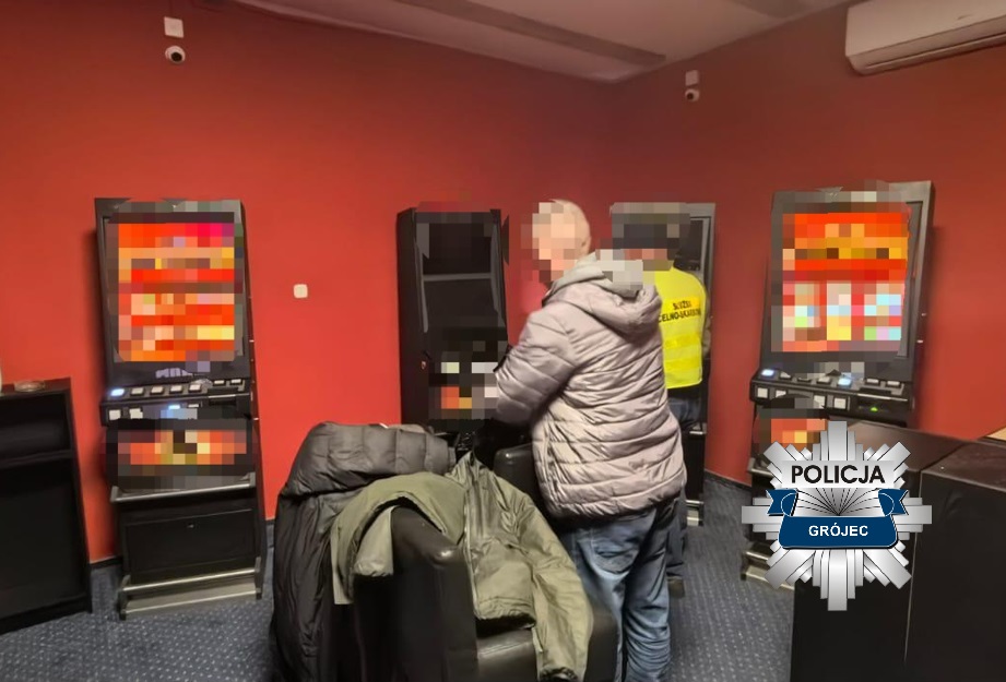 Mężczyzna w szarej kurtce obsługujący automat do gier w pomieszczeniu z czerwonymi ścianami, obok stoi pracownik ochrony.