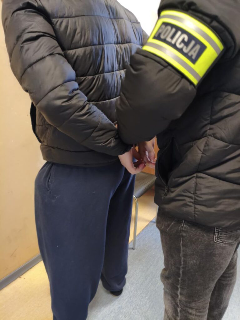 Mężczyzna w dresie jest zatrzymywany przez policjanta w mundurze w pomieszczeniu z jasnymi ścianami.