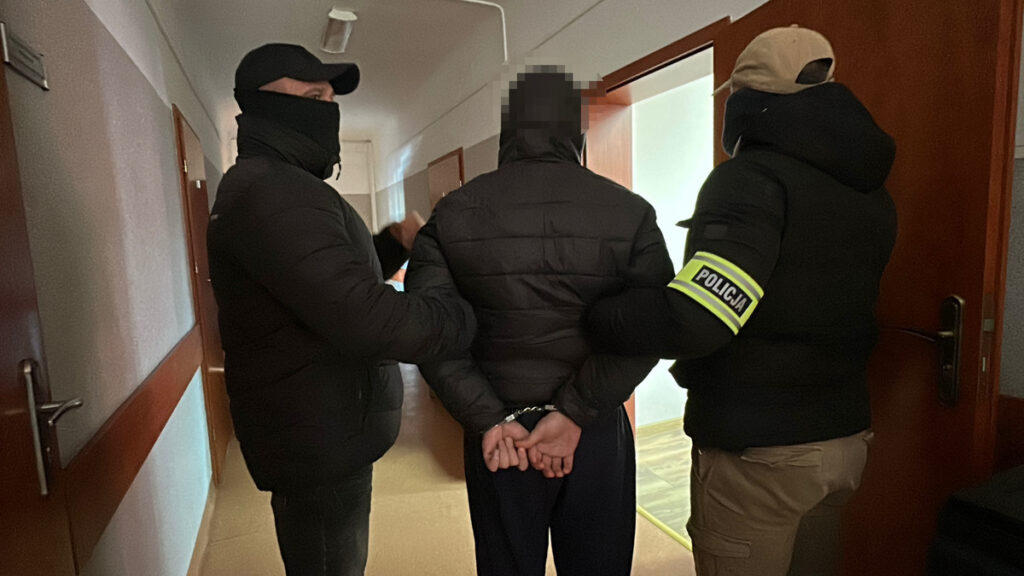 Trzech funkcjonariuszy w odzieży przystosowanej do działań operacyjnych eskortuje zatrzymanego mężczyznę w korytarzu budynku.