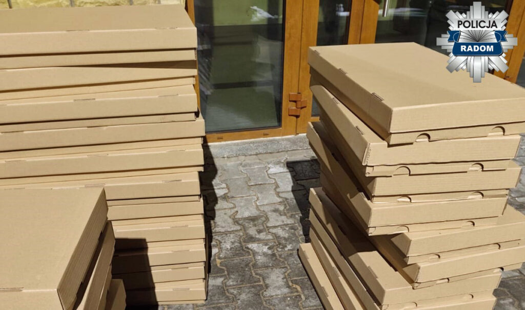 Stosy kartonowych pudełek leżą na chodniku przed dużymi przeszklonymi drzwiami.