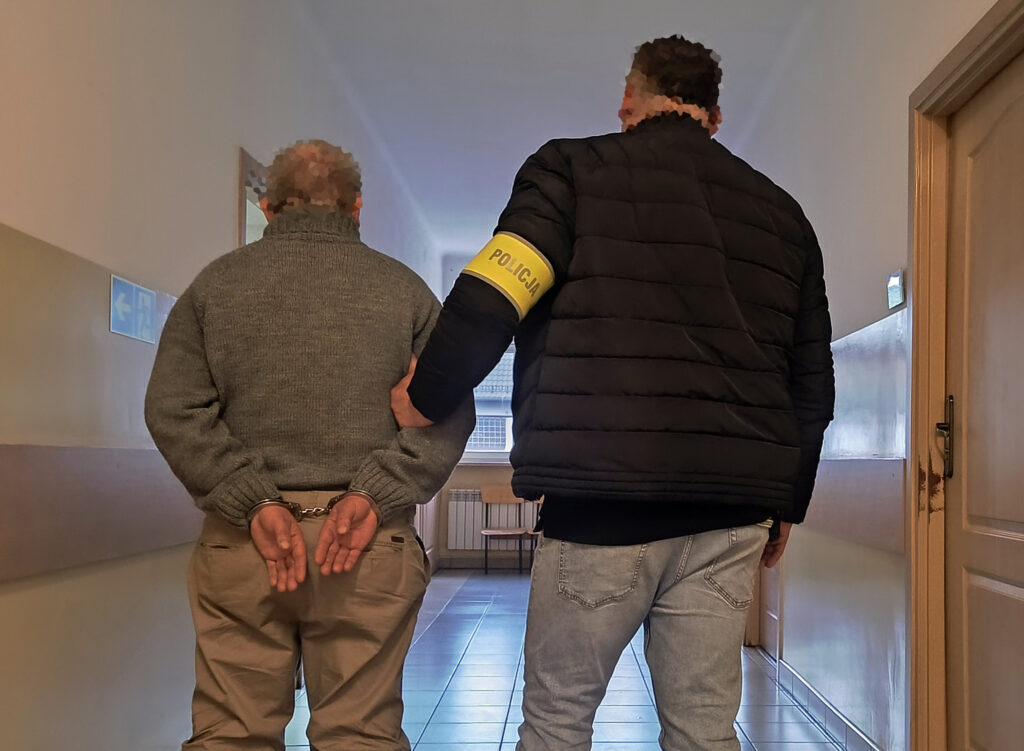 Mężczyzna w kajdankach prowadzony przez policjanta w czarnej kurtce w korytarzu budynku.