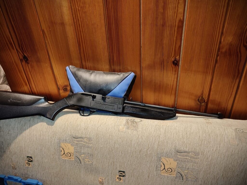 Karabinek pneumatyczny leży na kanapie, obok poduszki w niebiesko-czarnym kolorze, na tle drewnianej ściany.