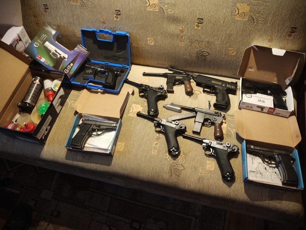 Na kanapie leżą różne pistolety w pudełkach, obok nich znajdują się butelki z kolorowymi kulkami oraz spray.