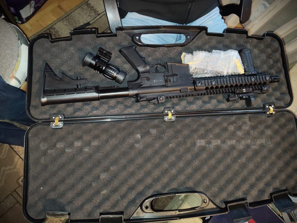 Karabin w etui, obok zamontowany celownik, na tle czarnej wyściółki.