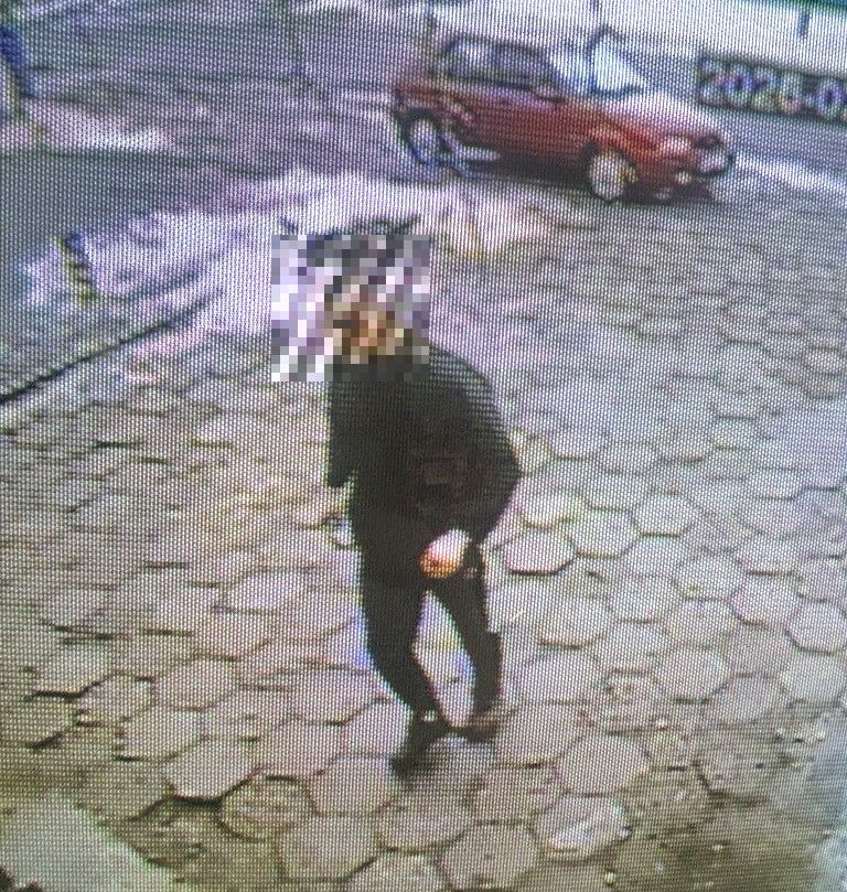 Mężczyzna w czarnym ubraniu idzie po kostkowanej nawierzchni, w tle widać czerwony samochód.