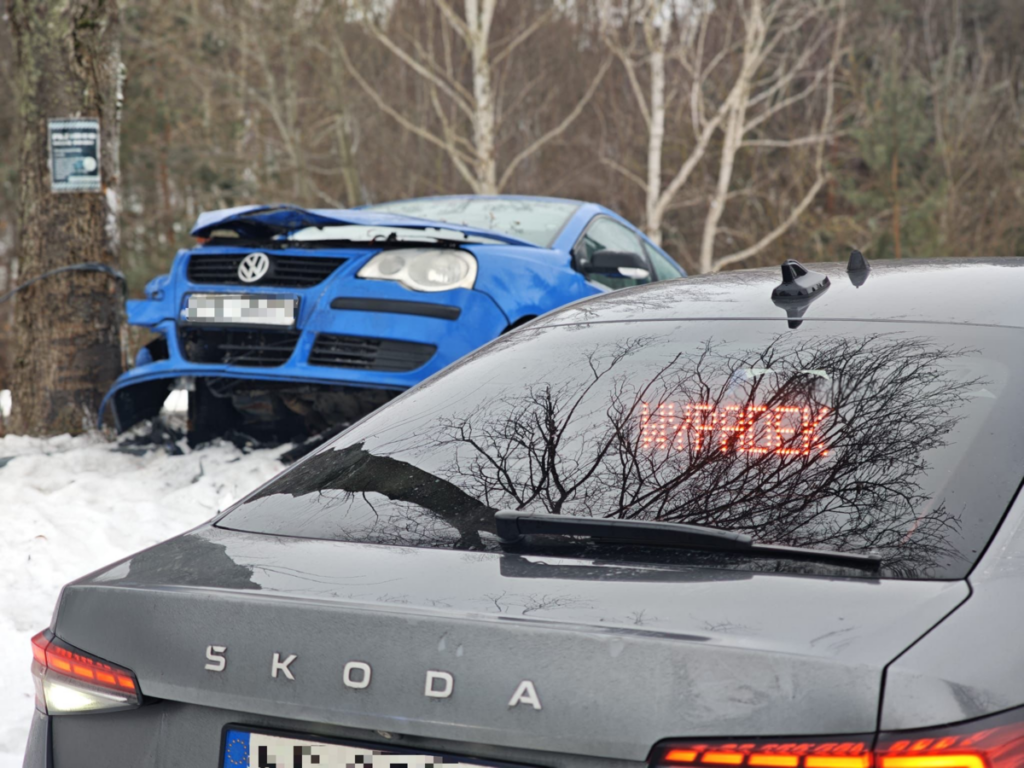 Niebieski samochód osobowy uderzony w drzewo, w tle widoczny czarny pojazd z wyświetlaczem LED. Scena w zimowym lesie.