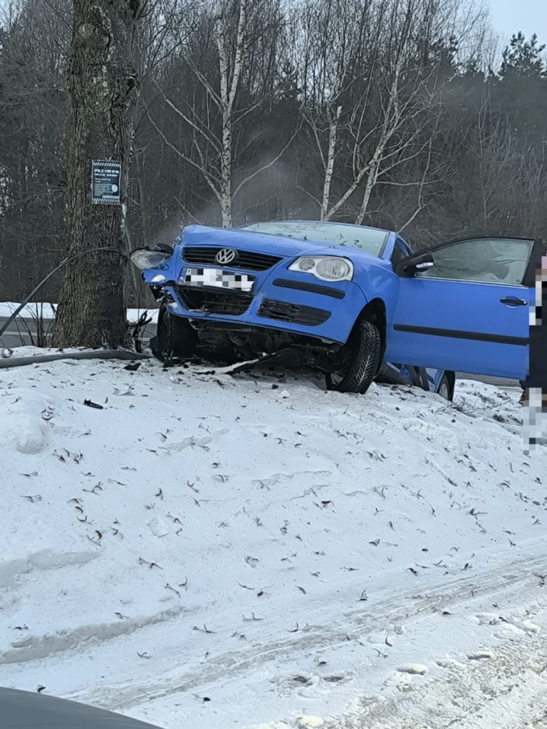 Niebieski samochód osobowy uderzył w drzewo na zaśnieżonej poboczu drogi, z otwartymi drzwiami po stronie kierowcy.