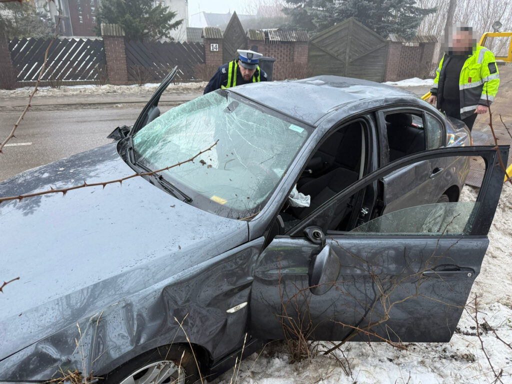 Policyjny funkcjonariusz bada uszkodzone auto osobowe z rozbitą szybą, obok stoi drugi mężczyzna w odblaskowej kurtce.