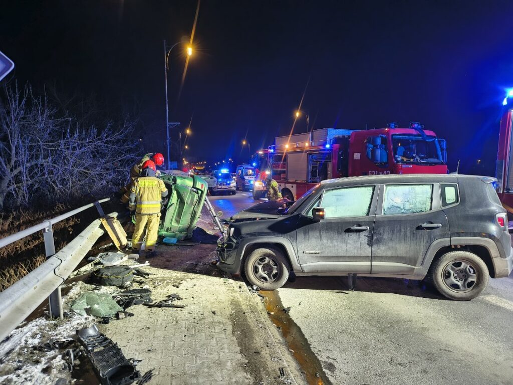 Strażacy w żółtych mundurach pomagają osobie z przewróconego samochodu na drodze, obok stoją inne pojazdy ratunkowe.