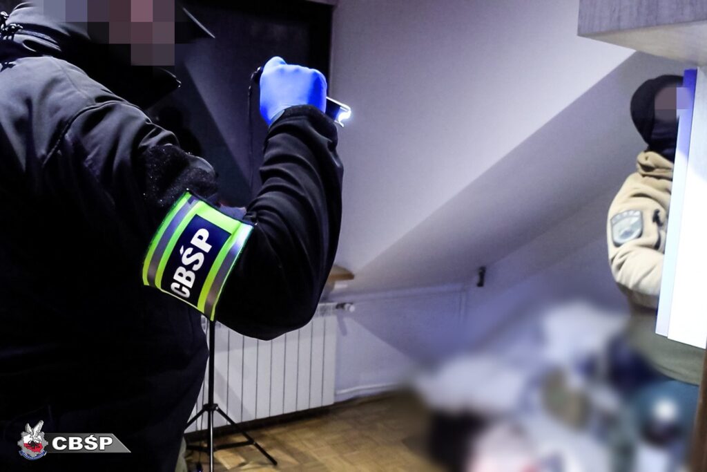 Funkcjonariusz CBŚP z latarką przeszukuje pomieszczenie, w tle widoczna postać w odzieży wierzchniej.