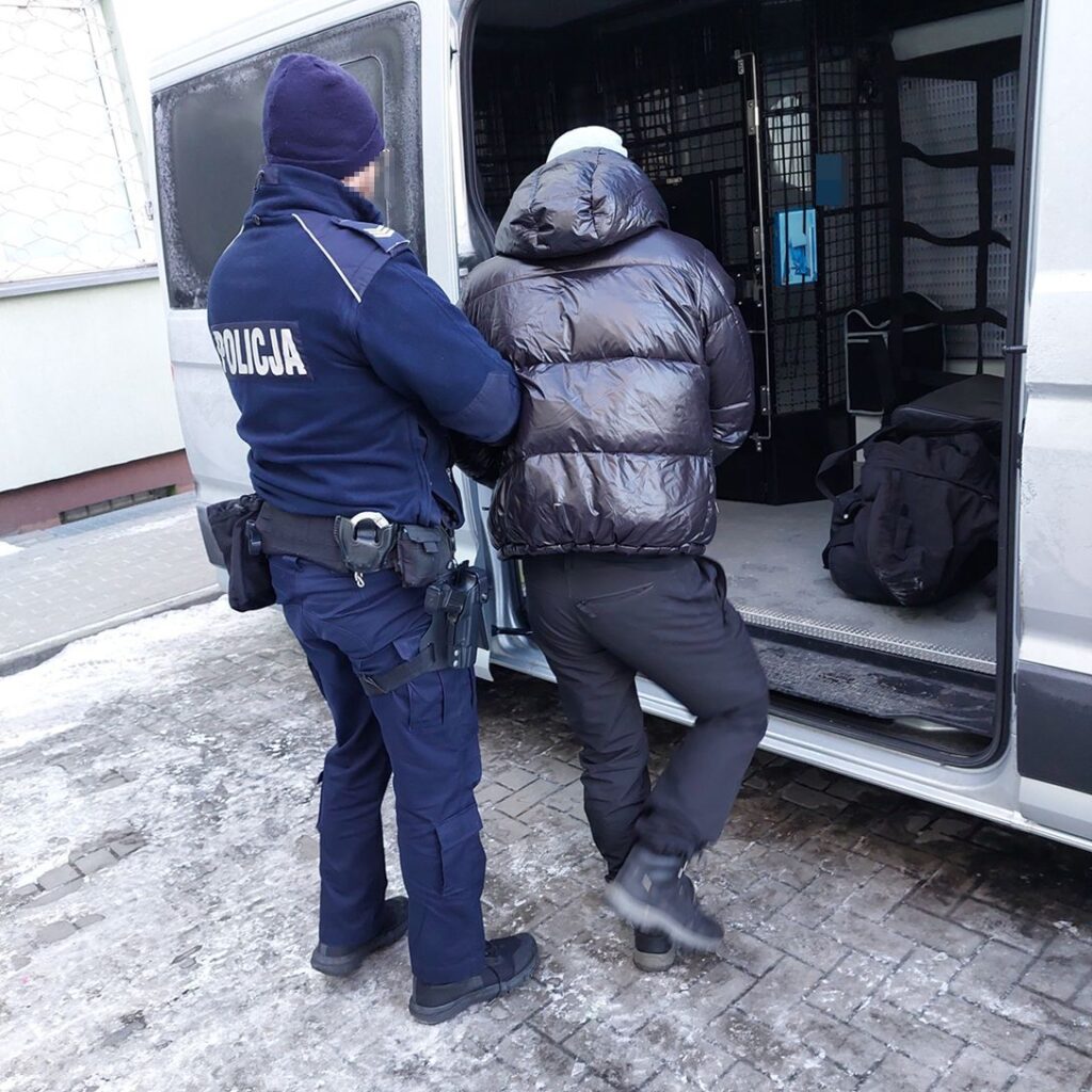 Policjant w mundurze prowadzi zatrzymanego mężczyznę w puchowej kurtce do policyjnego vana.