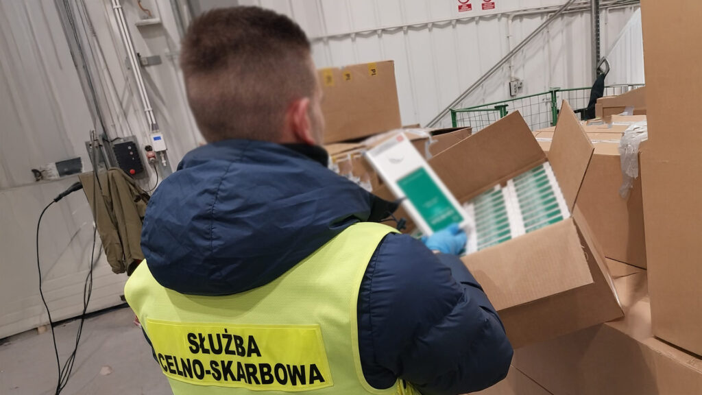 Funkcjonariusz służby celno-skarbowej sprawdza zawartość paczki w magazynie, trzymając w ręku dokumenty.