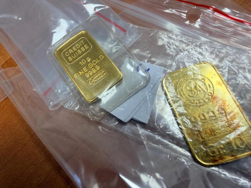 Dwie sztabki złota w przezroczystych opakowaniach na tle brązowego materiału.