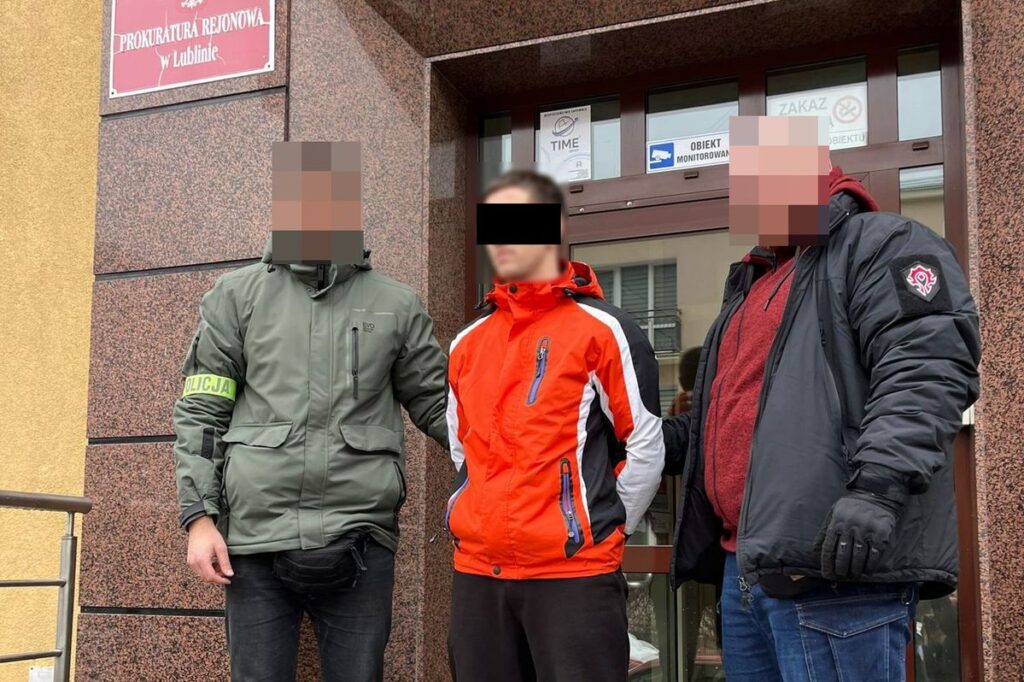 Mężczyzna w pomarańczowej kurtce stoi z dwiema osobami w mundurach przed budynkiem prokuratury.