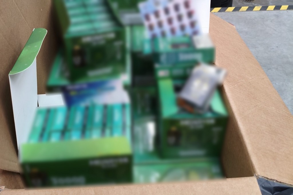 Pudełko wypełnione zielonymi opakowaniami z produktami, częściowo zasłonięte przez inne kartony.