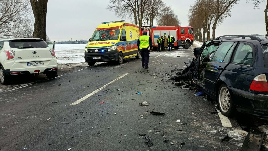 Na drodze leżą zniszczone elementy pojazdów, a obok zatrzymane są karetka i wóz strażacki; policjant kieruje ruchem.