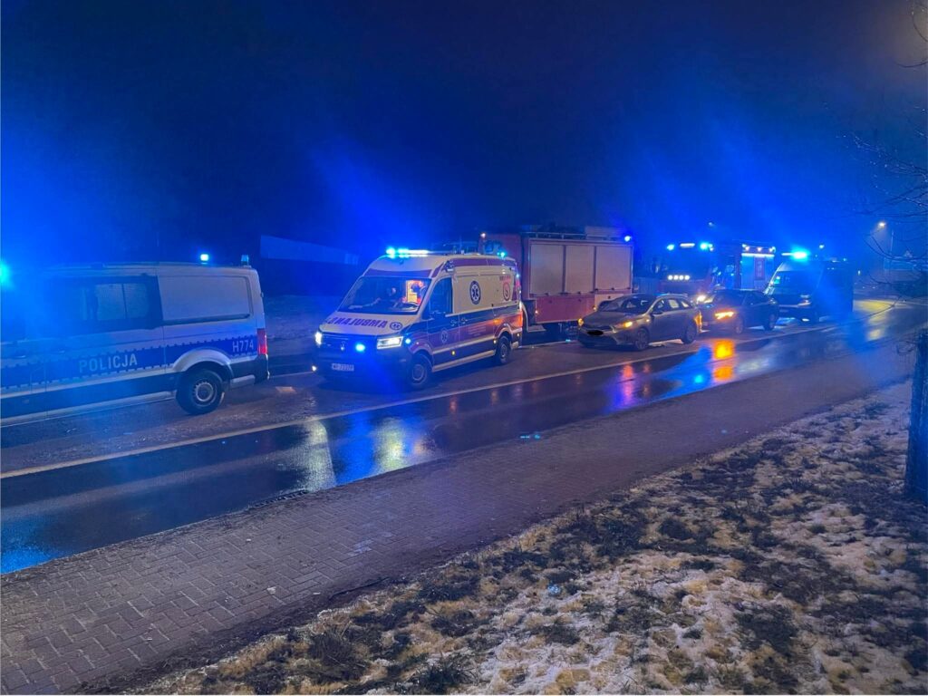 Na drodze stoją pojazdy służb ratunkowych z włączonymi sygnałami świetlnymi, otoczone przez mokrą nawierzchnię.