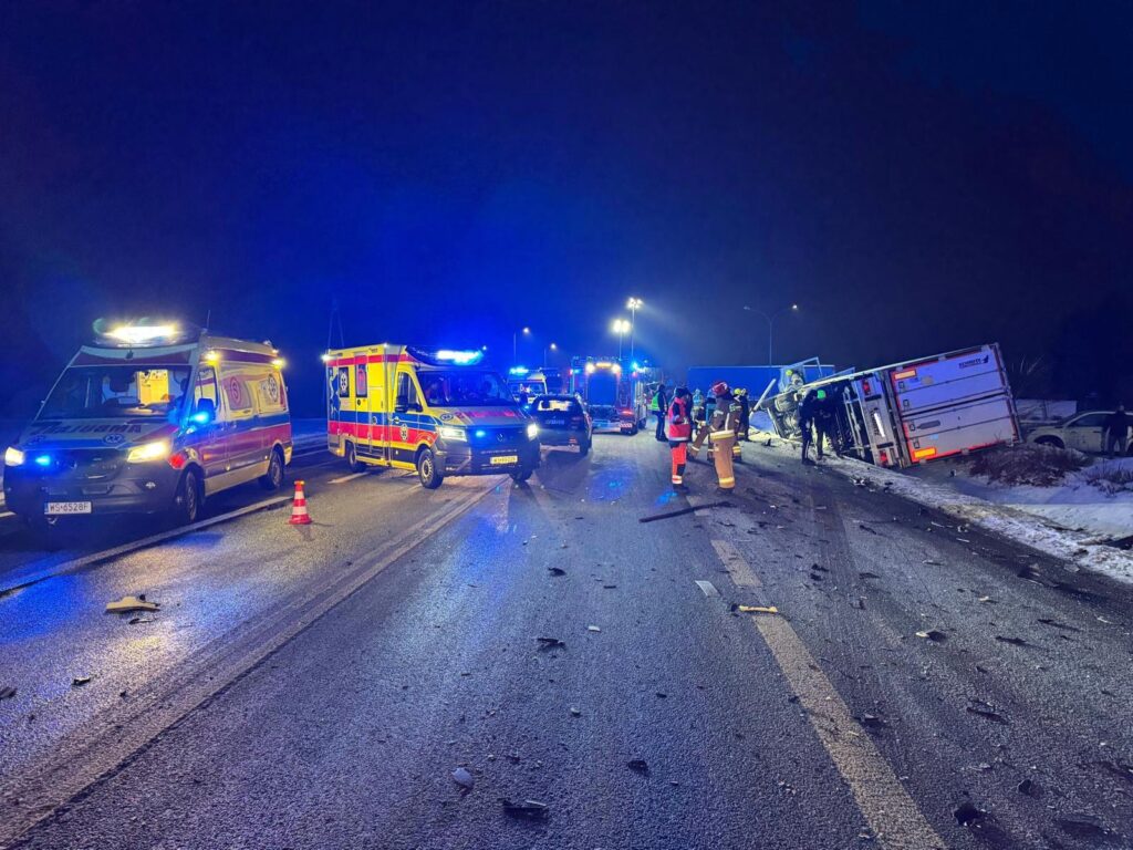 Na drodze nocą, zderzenie pojazdów; karetki i strażacy udzielają pomocy poszkodowanym przy przewróconym samochodzie.