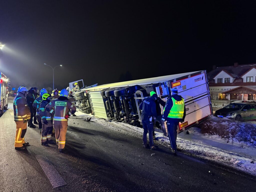 Grupa strażaków i policjantów bada przewrócony na bok ciężarówkę na ośnieżonej drodze nocą. W tle widoczny hotel.