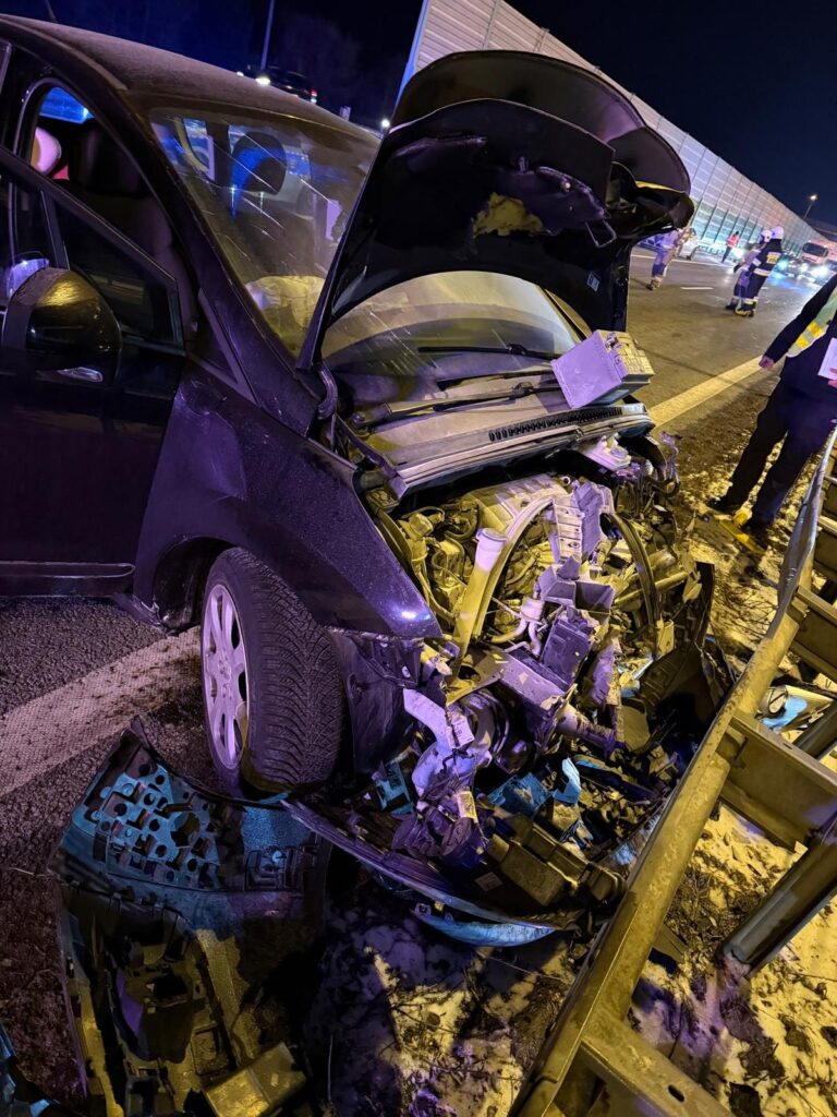 Uszkodzony samochód osobowy z otwartą maską stoi na poboczu drogi, obok zabezpieczeń, w nocy.