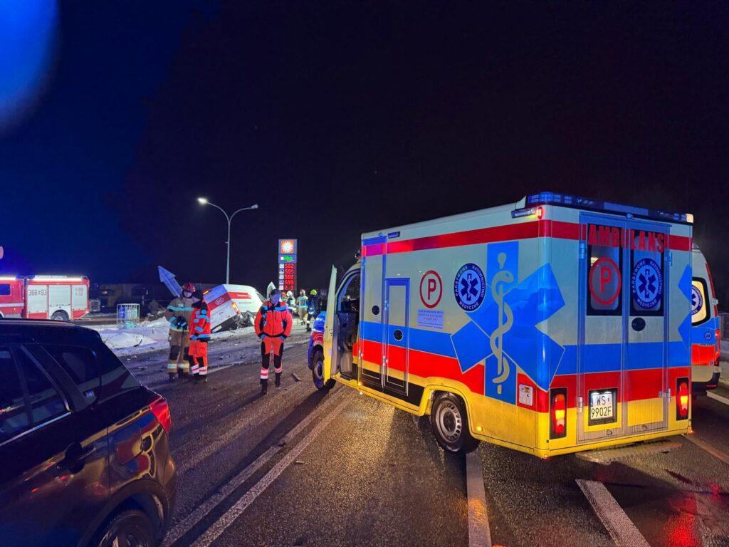 Ratownicy medyczni w odblaskowych strojach pracują przy wypadku drogowym obok przewróconego pojazdu w nocy.